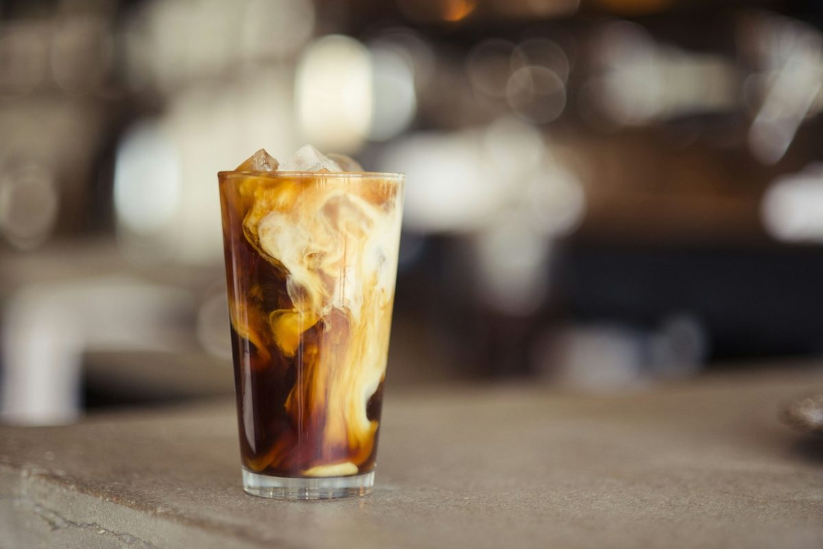 For 18feet Yonge and Dundas: A tall iced latte with cream marbling through it on a concrete counter, café lights blurred behind