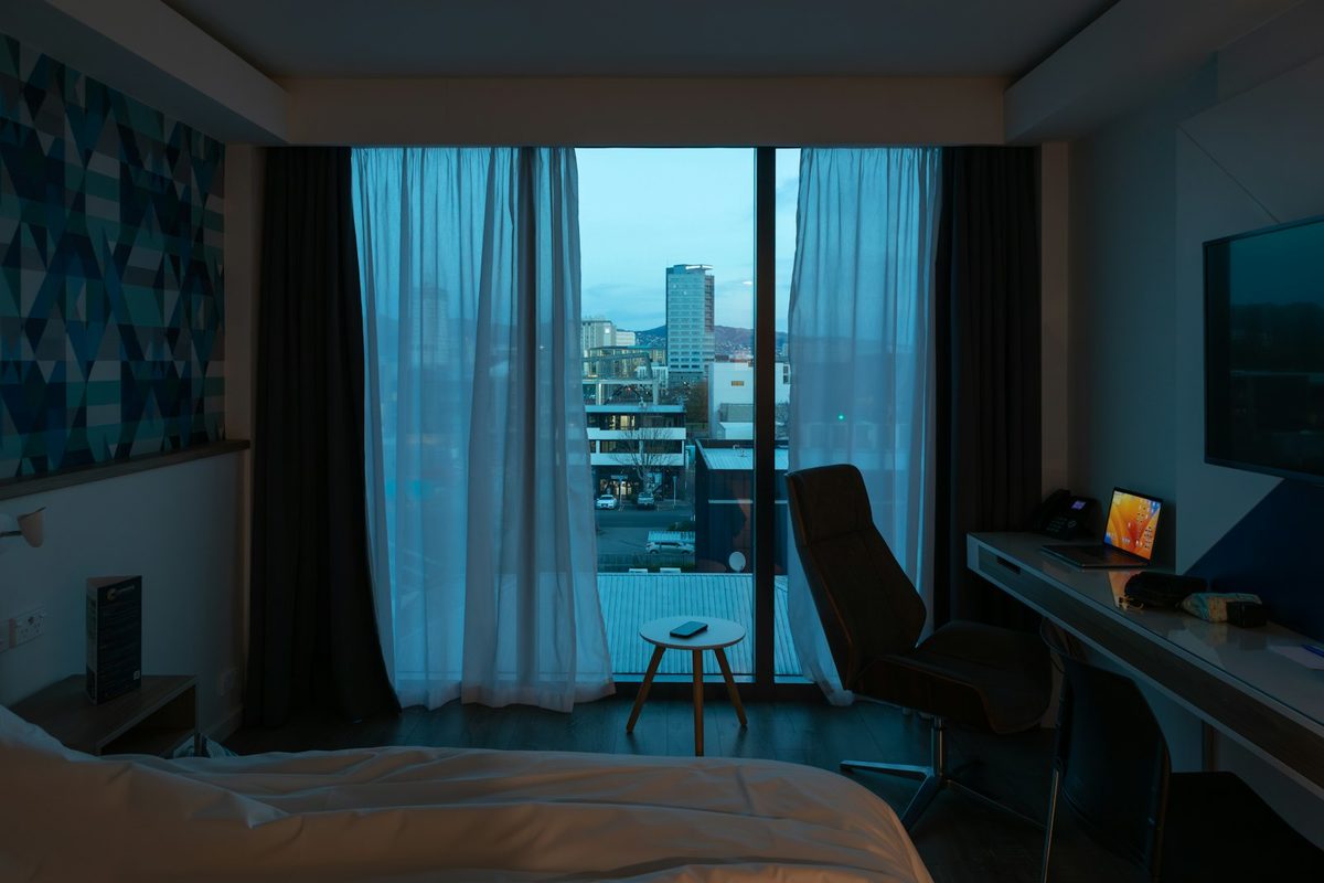 For Ace Hotel Toronto: A hotel room at dusk with a desk and chair, sheer curtains half open on a city view