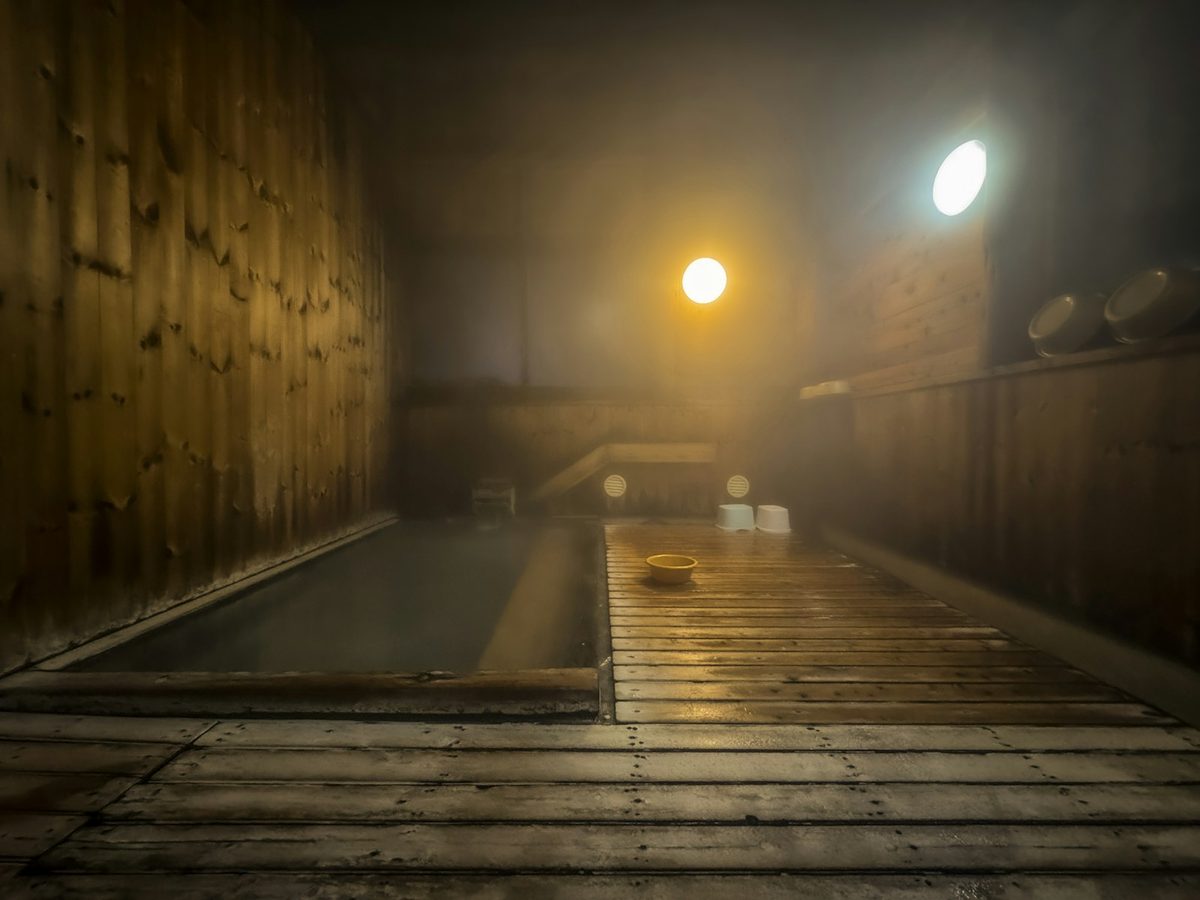 For AIRE Ancient Baths: A misty wooden bathhouse with a long sunken bath and two soft lamps glowing through the steam