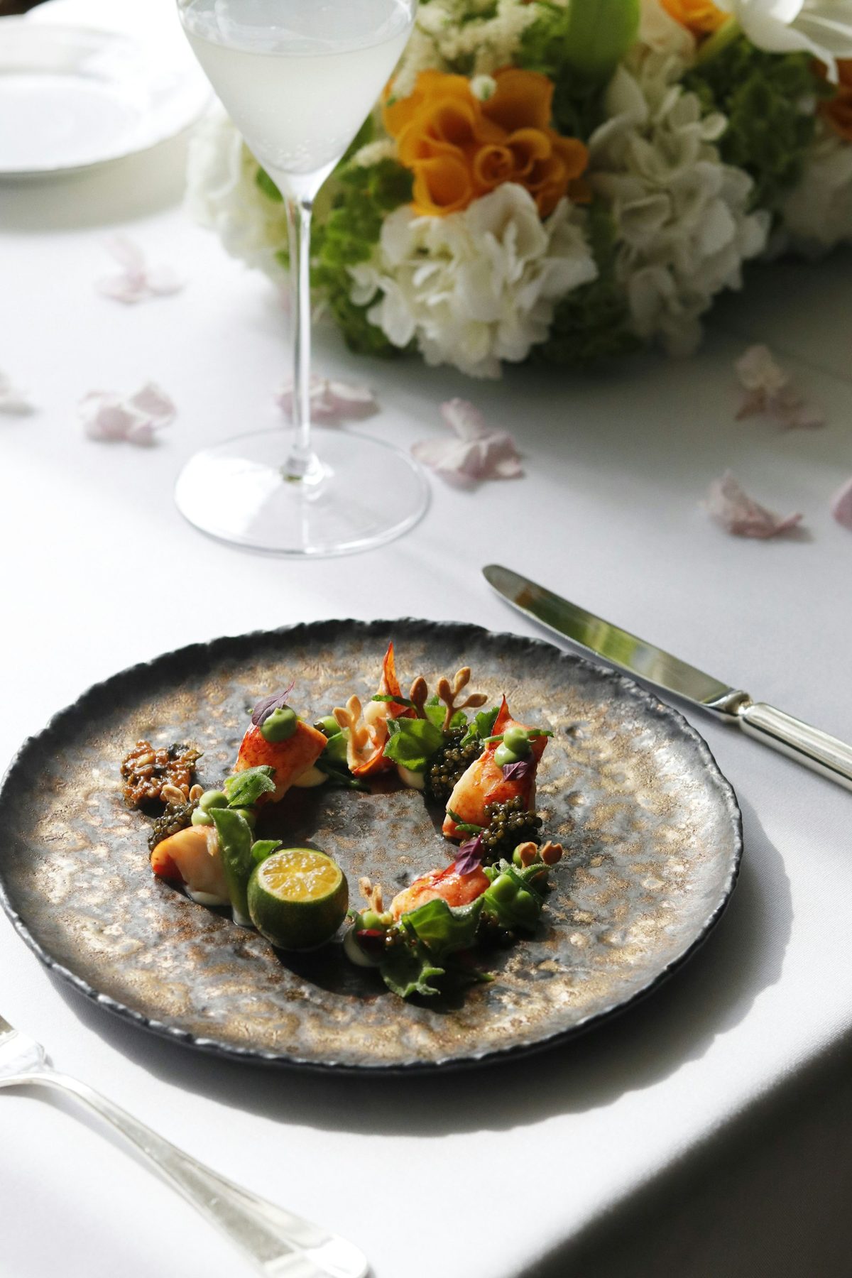 For Alo: A small composed course on a dark plate on a white tablecloth, wine glass and flowers behind