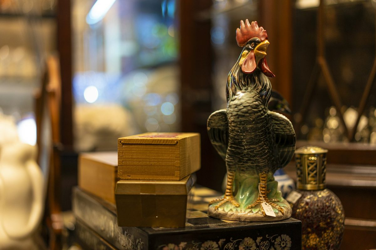 For Avenue Daughter: A painted ceramic rooster beside two wooden boxes and a small enamelled bottle on a patterned chest, warm shop lights blurred behind
