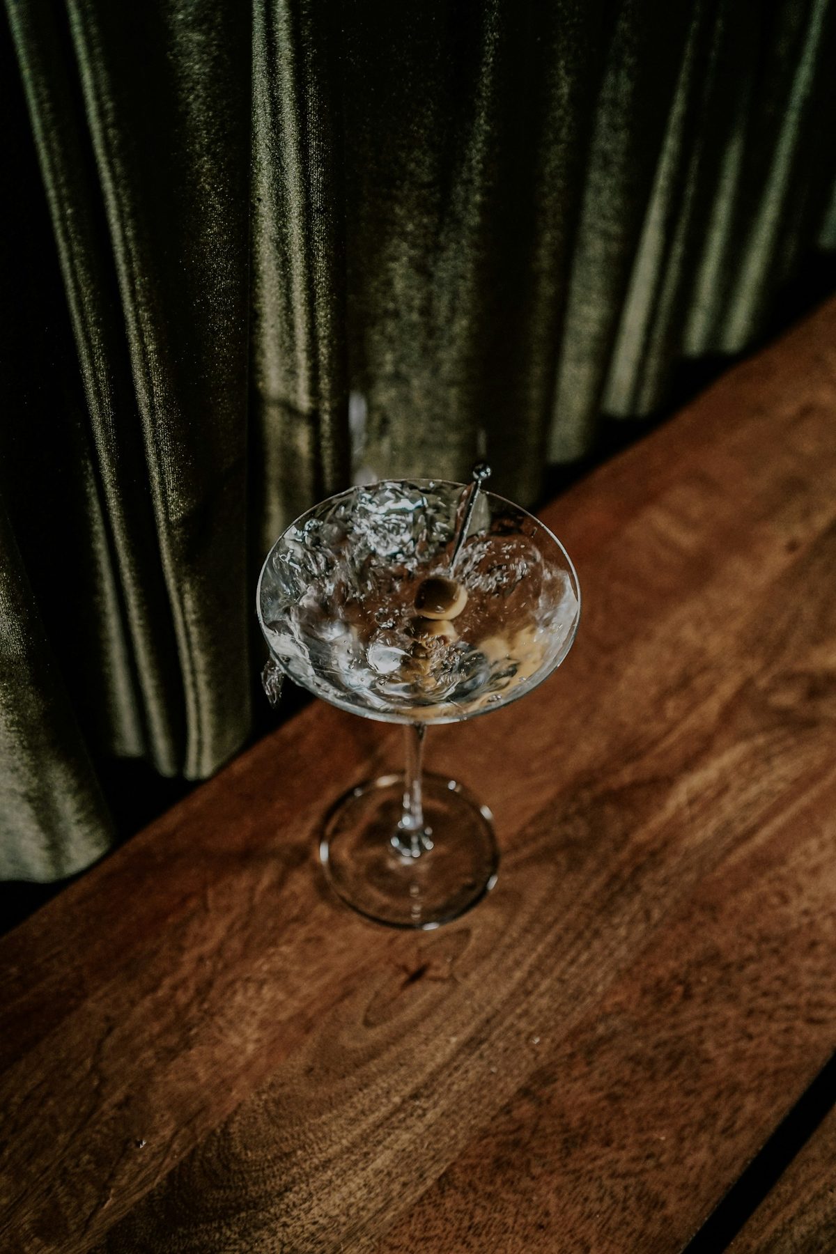 For Bar Pompette: A martini with olives on a dark wooden table beside a heavy green curtain