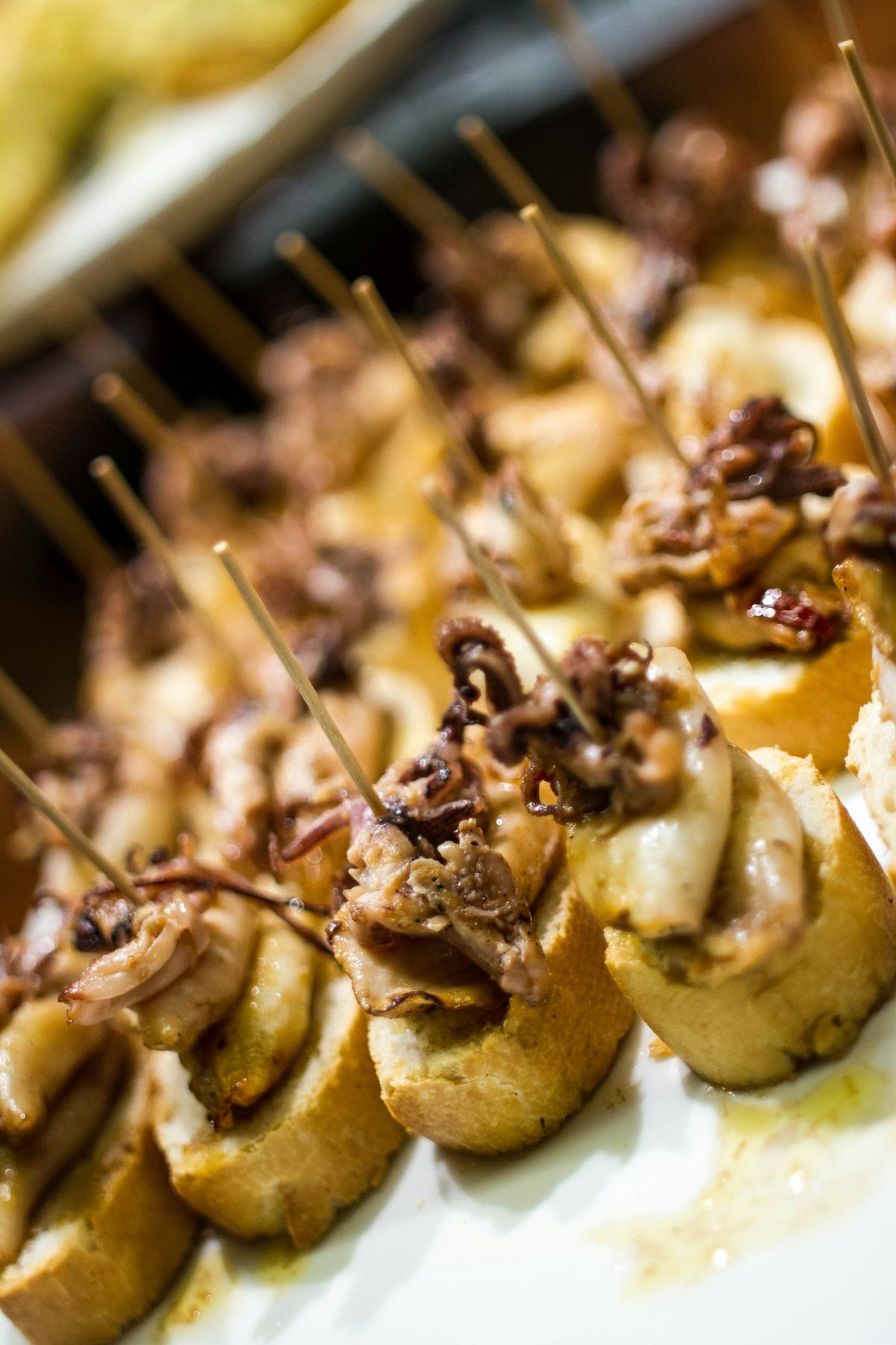 For Bar Raval: Close, shallow-focus row of pintxos on bread with toothpicks, warm light