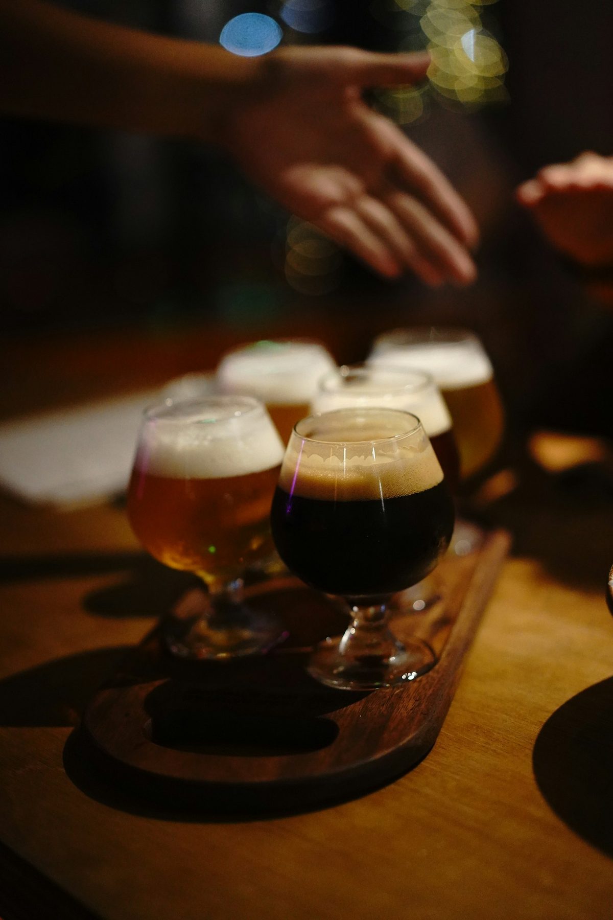For Bellwoods Brewery: A flight of dark and amber beers in stemmed glasses on a wooden paddle, hands reaching in, warm low light
