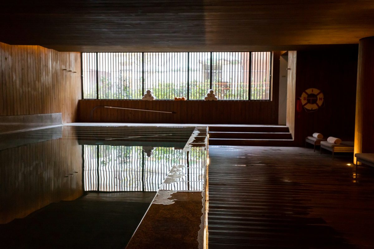 For Body Blitz Spa: A dim indoor pool with wooden walls, a long window at the far end reflected in the water