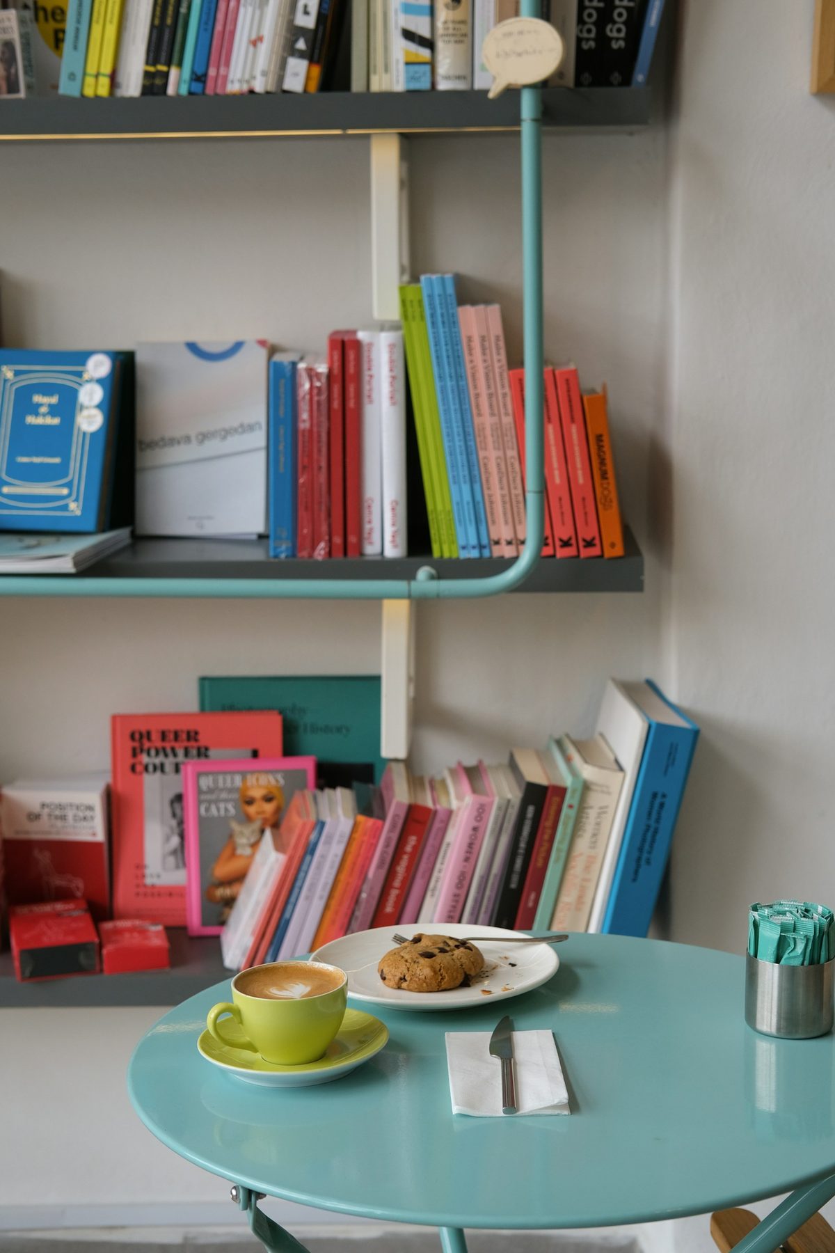 For Book Bar: A latte in a lime-green cup and a chocolate chip cookie on a small teal café table in front of shelves of colourful paperbacks (portrait)
