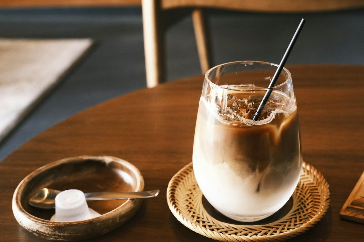 For Botanic Coffee & Caphe: An iced coffee with cream swirling through it on a woven rattan coaster, on a round wooden table in soft daylight.