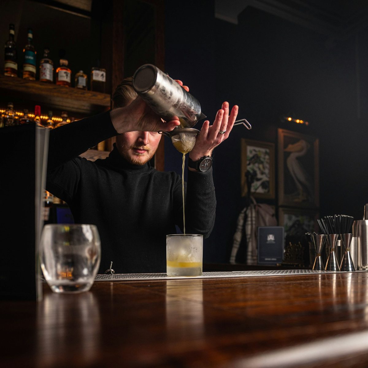 For Civil Liberties: A bartender straining a shaker into a glass at a dark wooden bar, shelves of bottles glowing behind