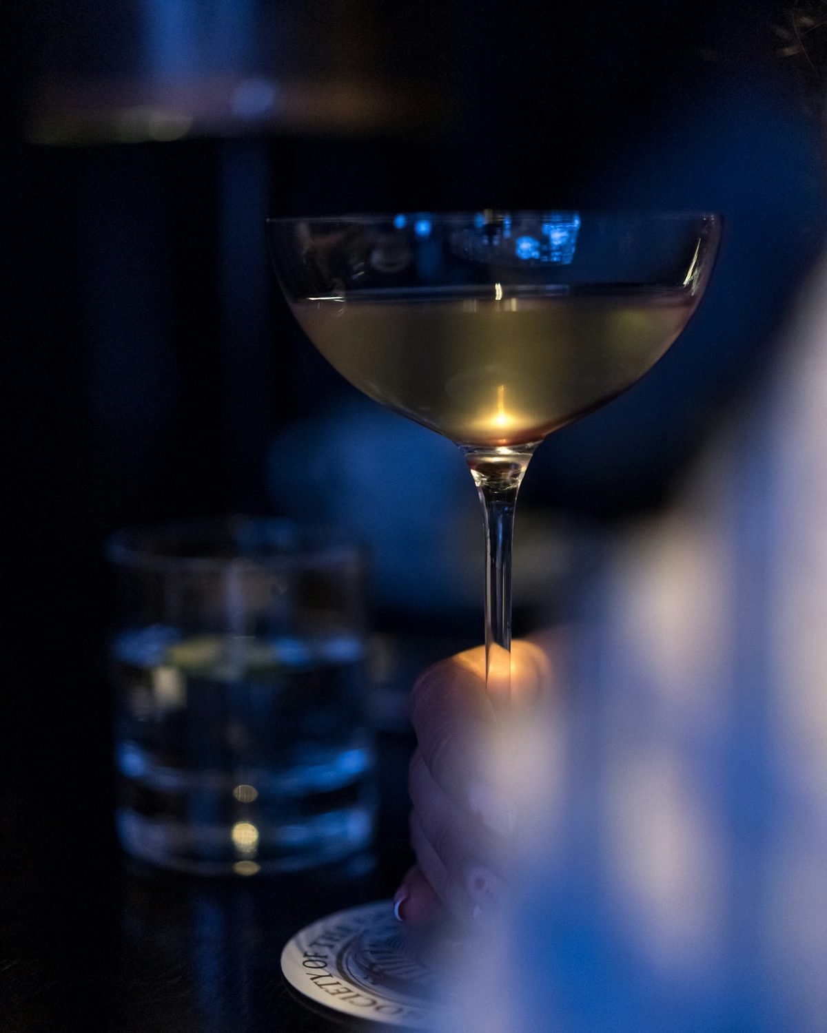 For Cry Baby Gallery: A coupe of pale cocktail held at its foot, lit from below in deep blue darkness, a water glass blurred behind
