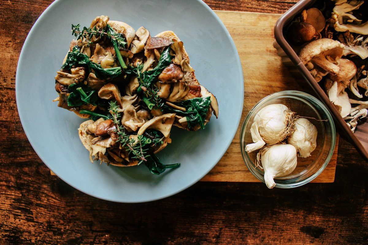 For Edulis: Overhead of sautéed wild mushrooms on toast on a pale plate, raw mushrooms and garlic on a dark wooden board