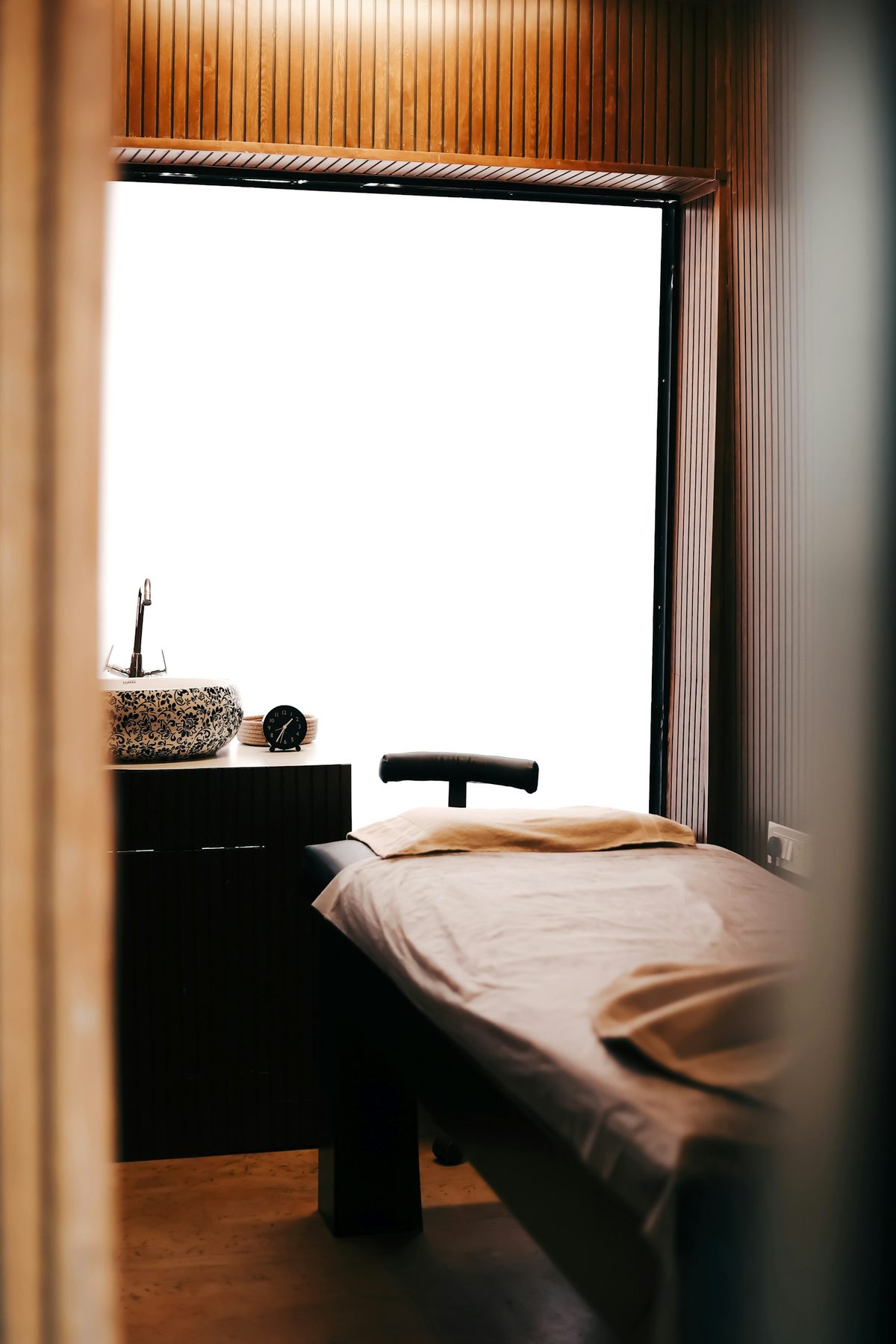 For Elmwood Spa: A quiet treatment room: a massage table in a beige sheet by a tall bright window, a patterned stone basin