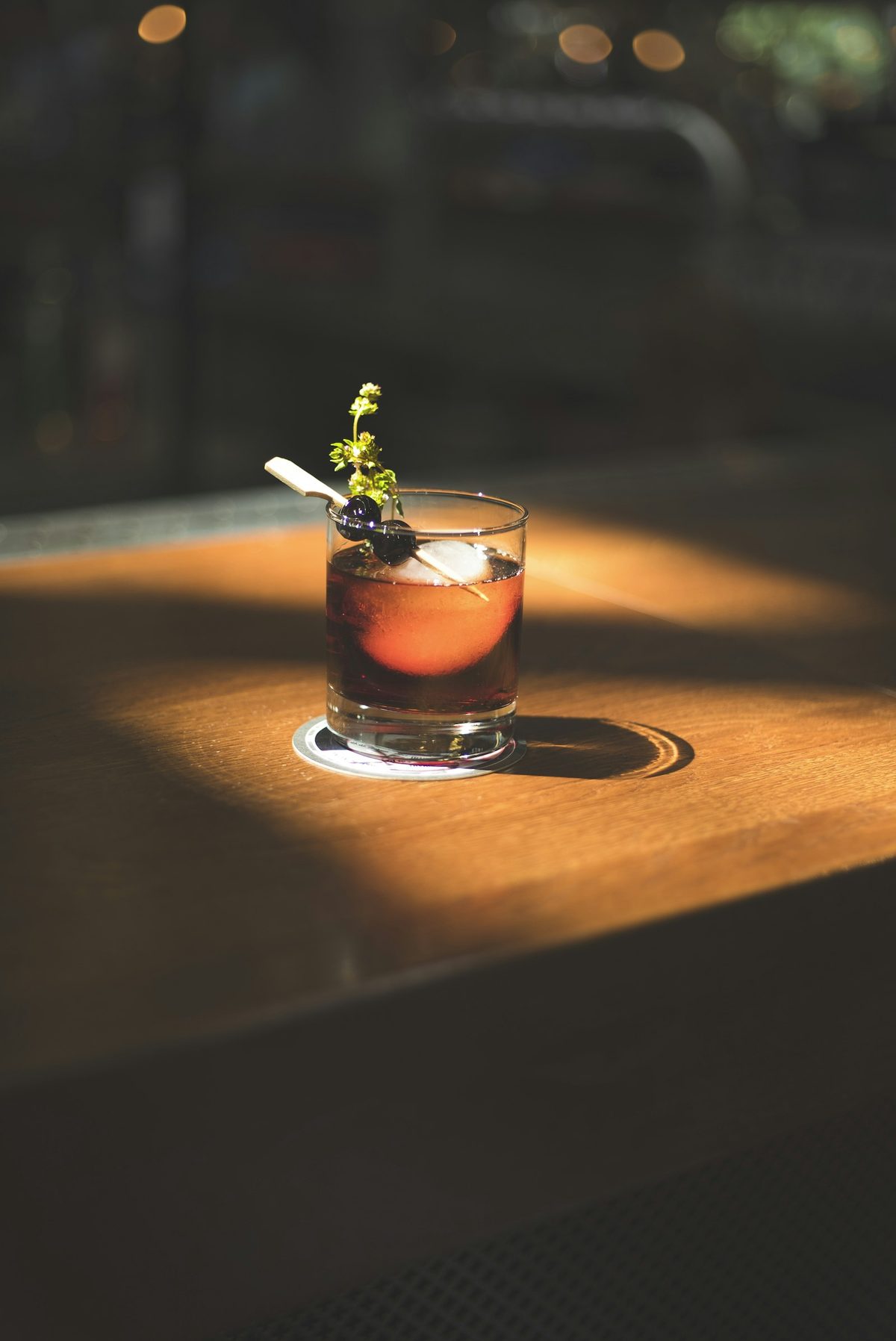 For Kensei Bar: A dark cocktail with an herb garnish on a wooden bar top in a pool of warm light