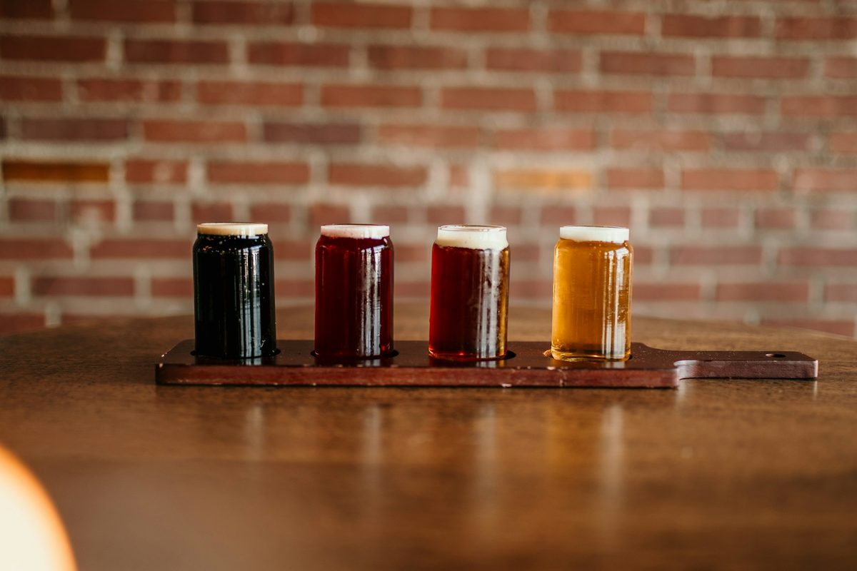 For Left Field Brewery: A flight of four beers, dark to golden, on a wooden paddle on a bar against a red brick wall