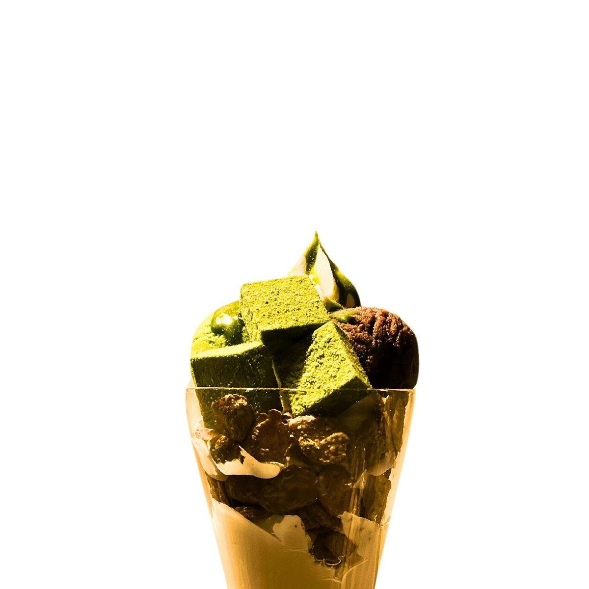 For Lost in Tokyo: A tall matcha parfait topped with matcha cake cubes and soft serve over chocolate layers, on a plain white background