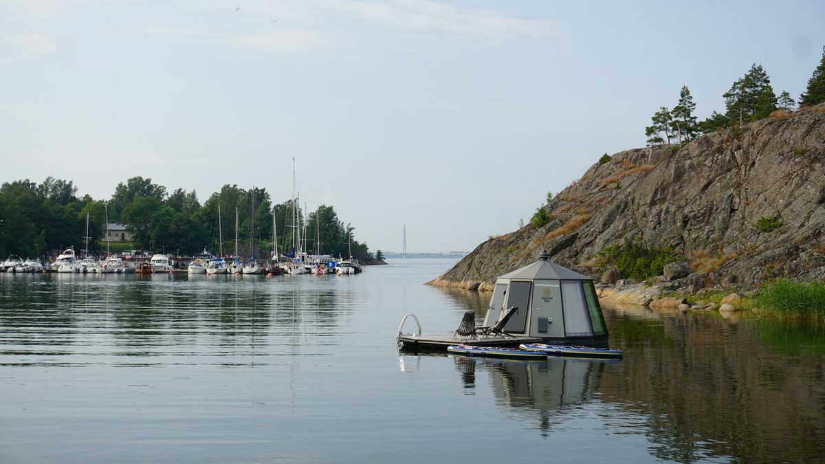 For Löyly Floating Sauna: A small floating sauna cabin moored in still water beside a rocky shore, boats far behind