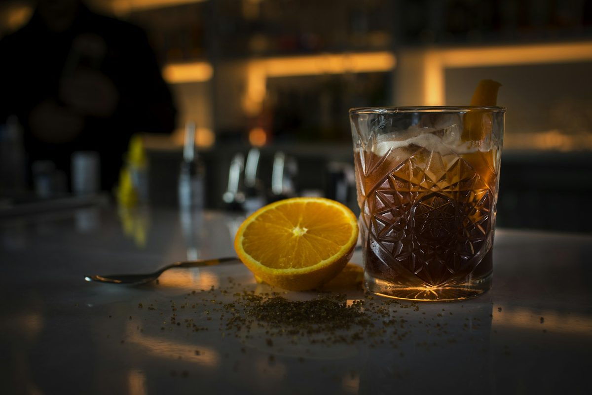 For Mother: A dark cocktail in a cut-glass tumbler with an orange half beside it on a bar, warm blurred lights behind