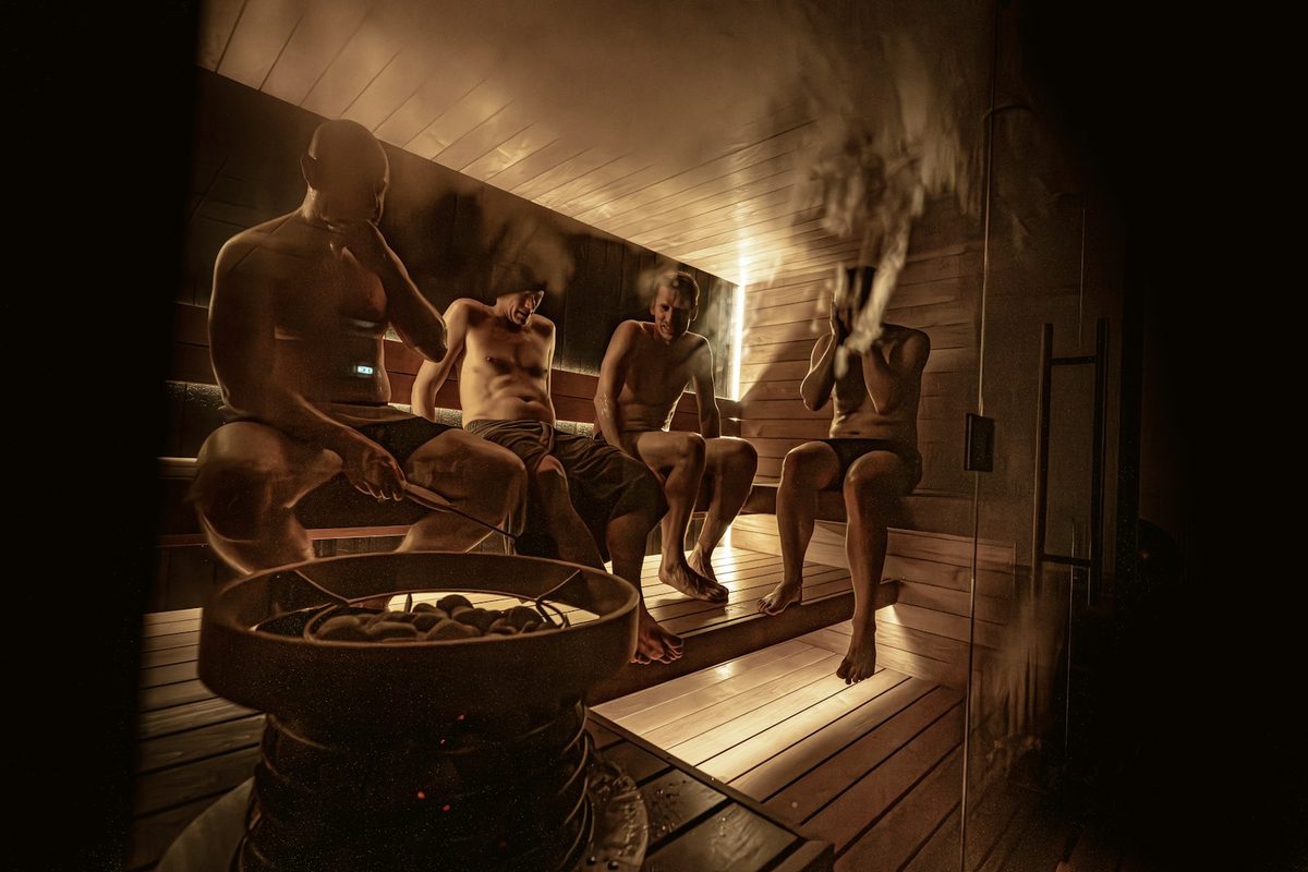 For Othership Adelaide: Four people sitting in a dim wooden sauna around a stone heater, steam rising