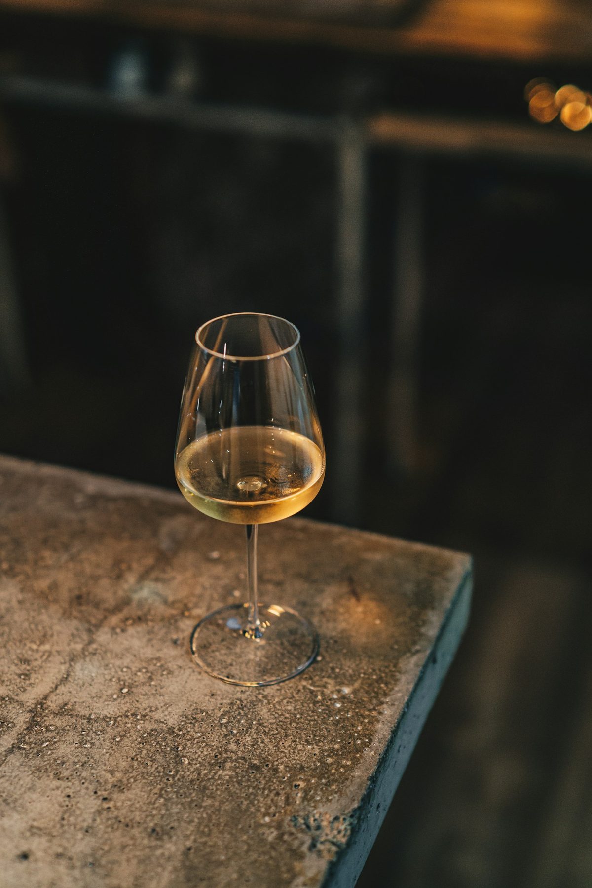 For Paradise Grapevine: A single glass of pale golden wine on a rough stone ledge in a dim room