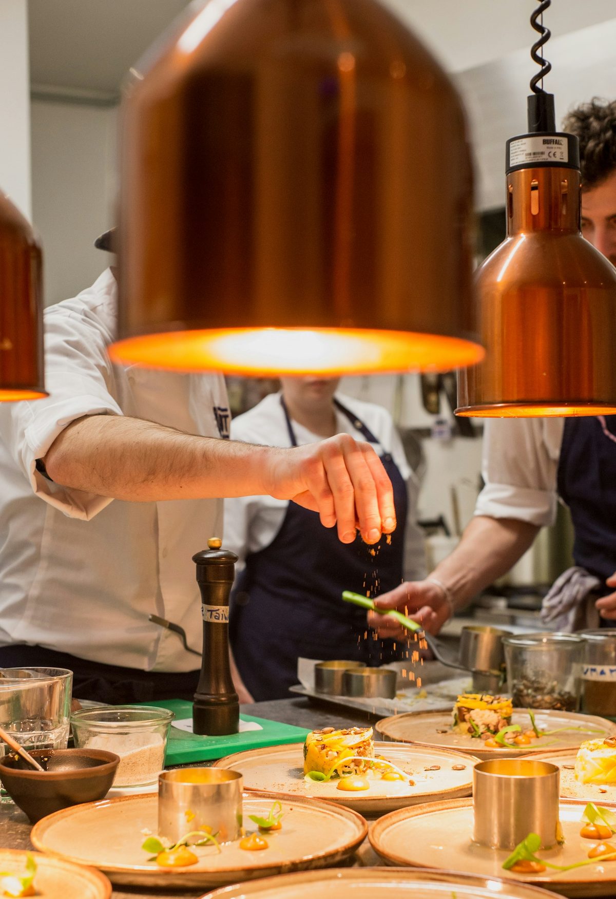 For Restaurant 20 Victoria: Cooks plating a row of small dishes under copper heat lamps at a kitchen pass