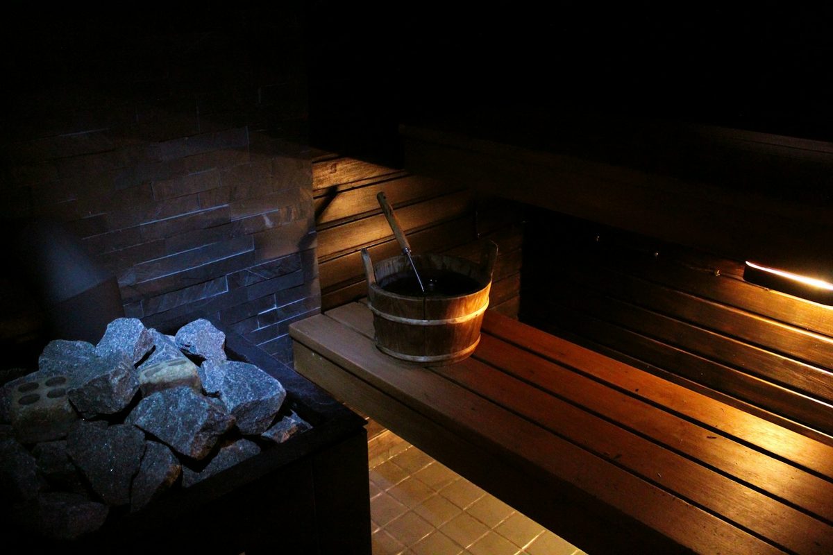 For SANA: Hot sauna stones in the foreground, a wooden bucket and ladle on a softly lit bench behind
