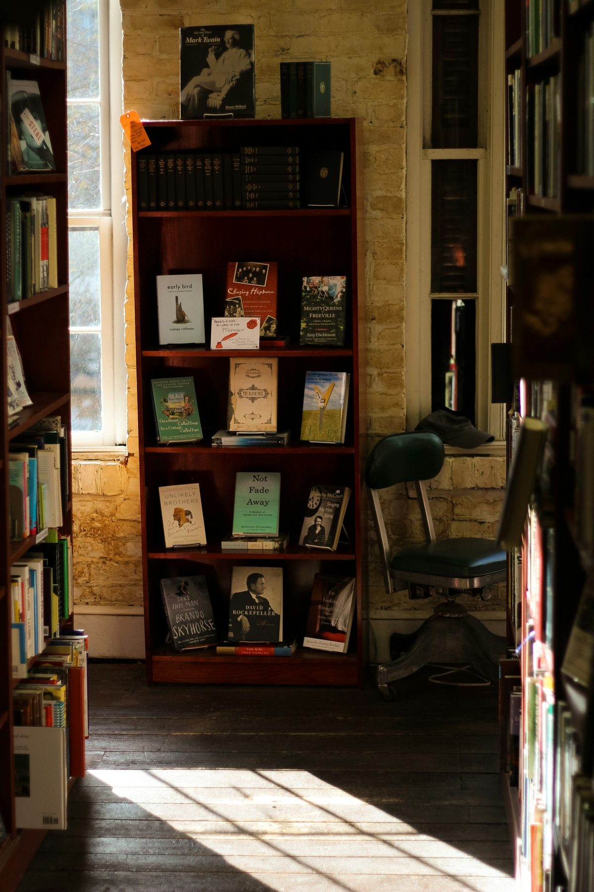 For She Said Boom!: A sunlit corner of a used bookshop: a wooden case of face-out books against a pale brick wall, a window and an old office chair, light across the floorboards
