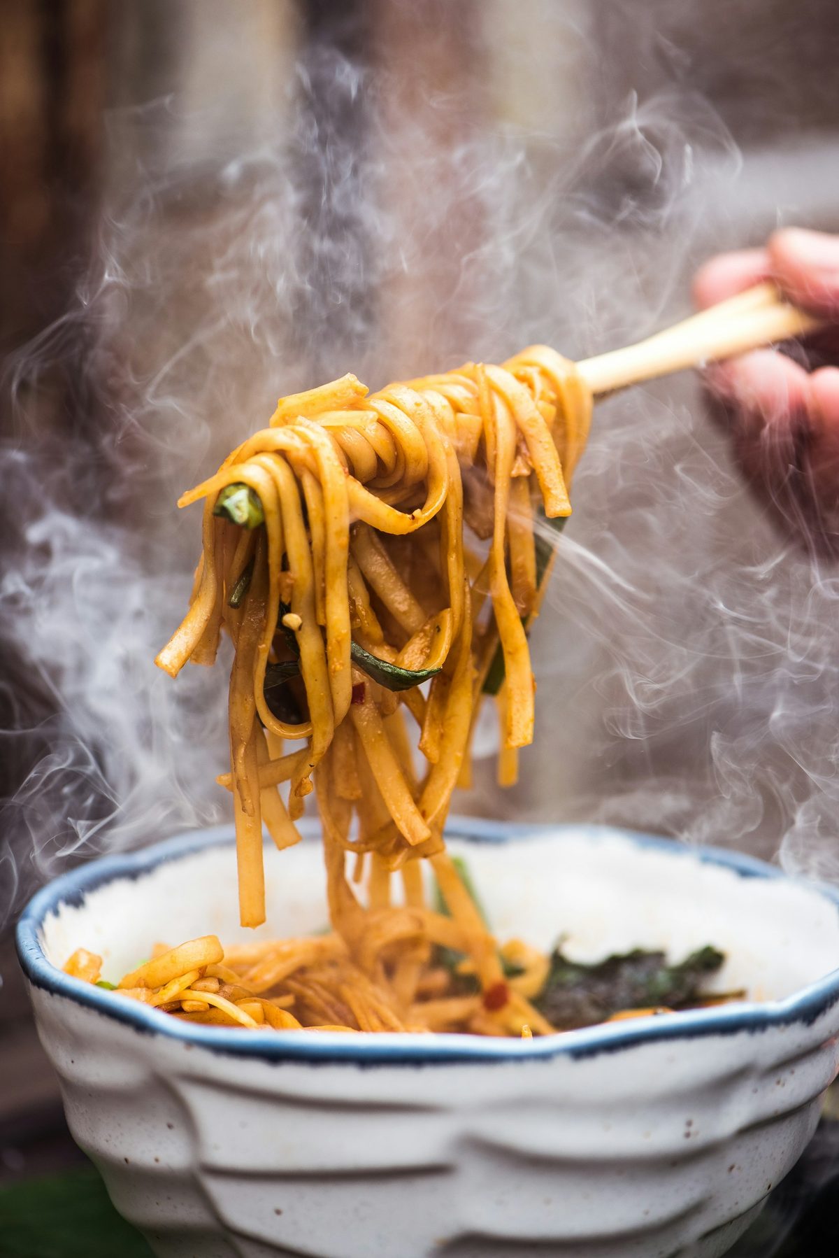 For Sunnys Chinese: Thick noodles lifted with chopsticks from a steaming patterned bowl, dark background
