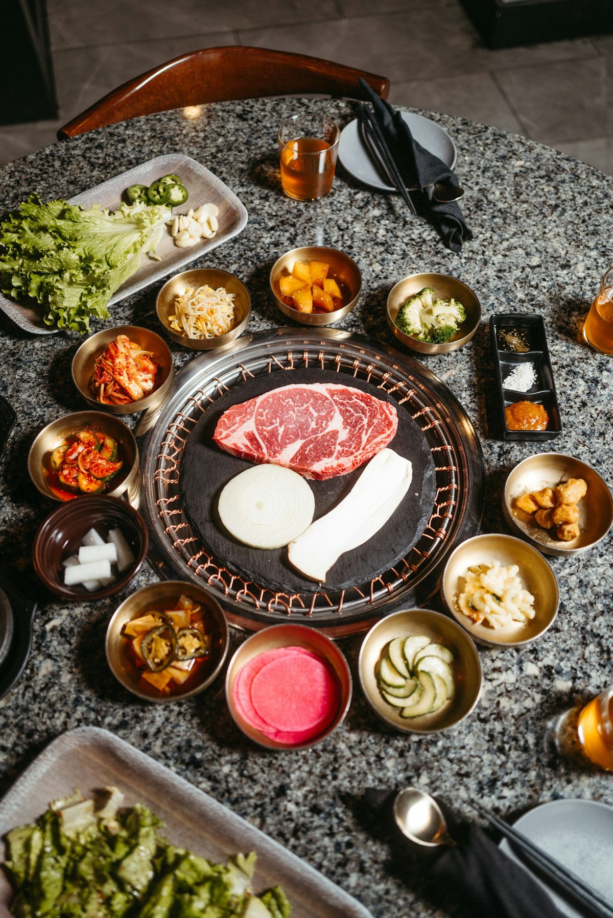 For Takja BBQ House: Overhead of a Korean barbecue table: marbled beef on the grill plate ringed by small banchan dishes