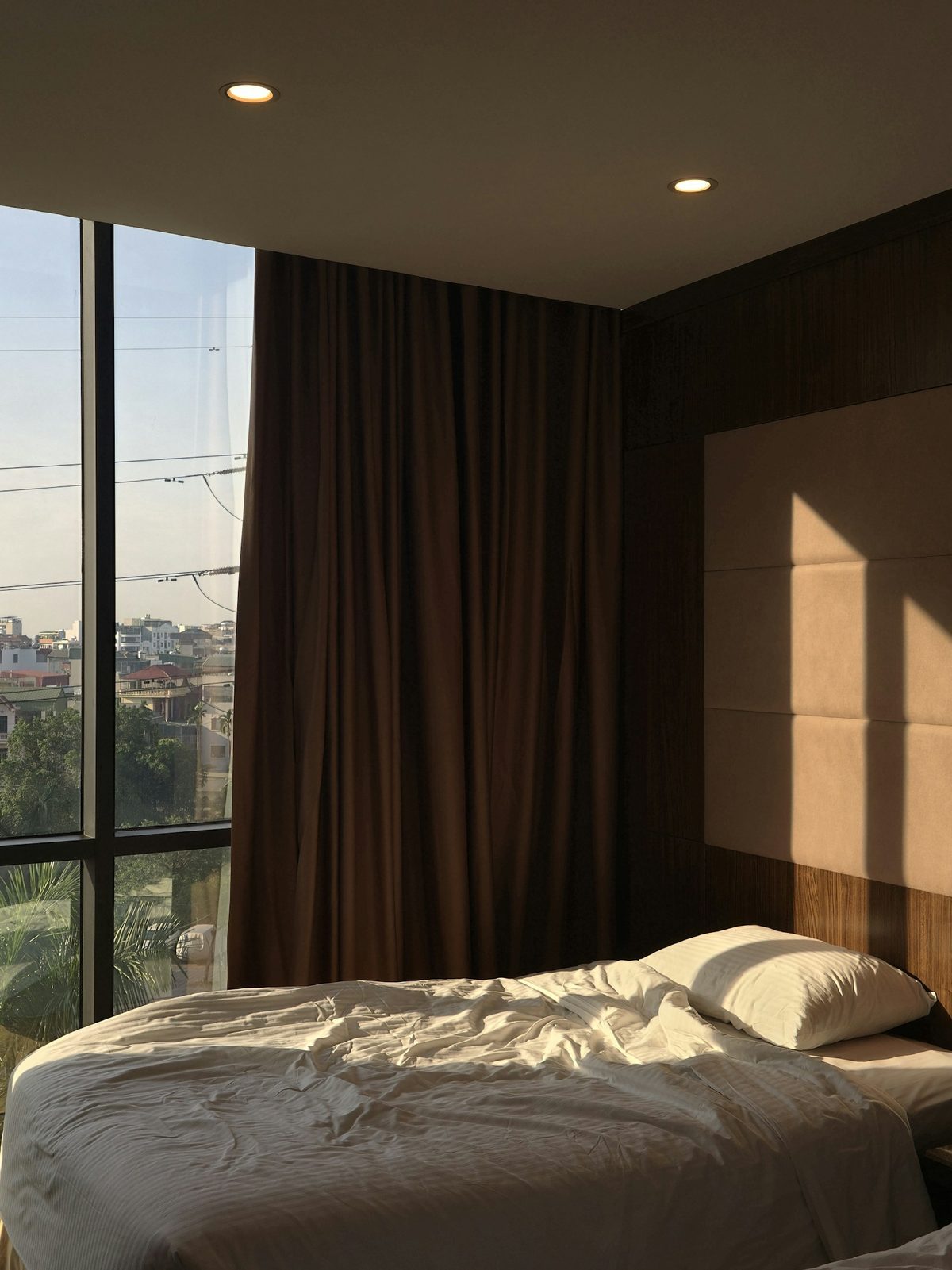 For The Hazelton Hotel: Morning sun through floor-to-ceiling windows across an unmade white bed, dark wood and heavy curtains