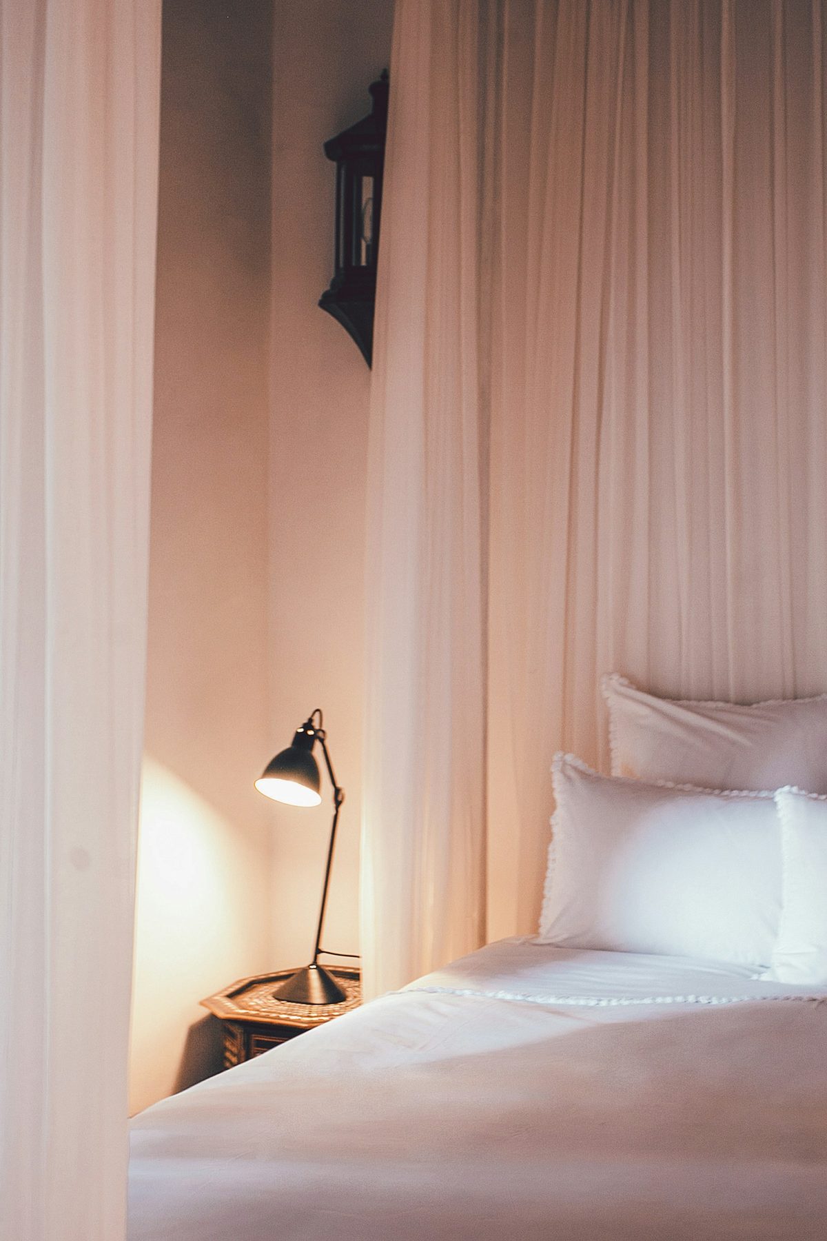 For The Ivy at Verity: A white bed and a black reading lamp in soft pink light through sheer curtains