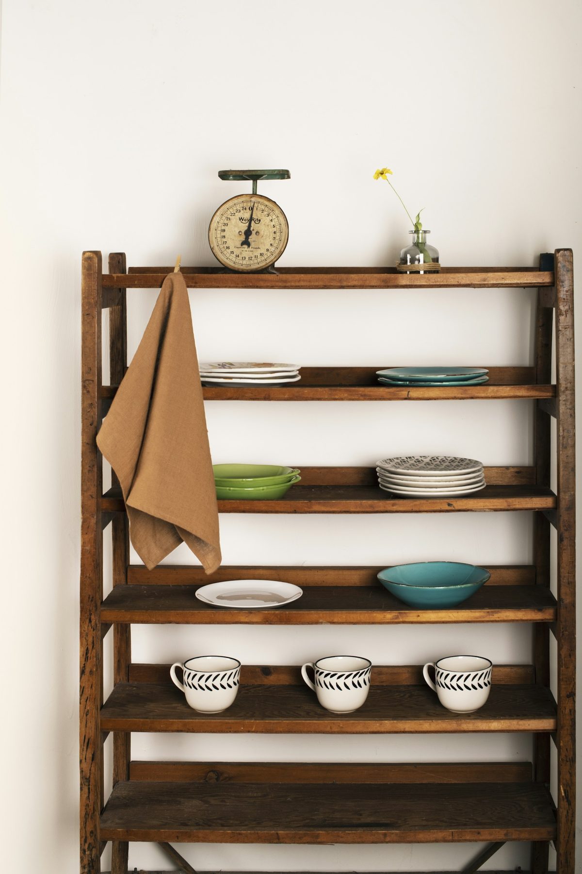For Tiny's General Store: An old wooden shelf against a white wall holding plates, three patterned cups, a tea towel and a vintage kitchen scale